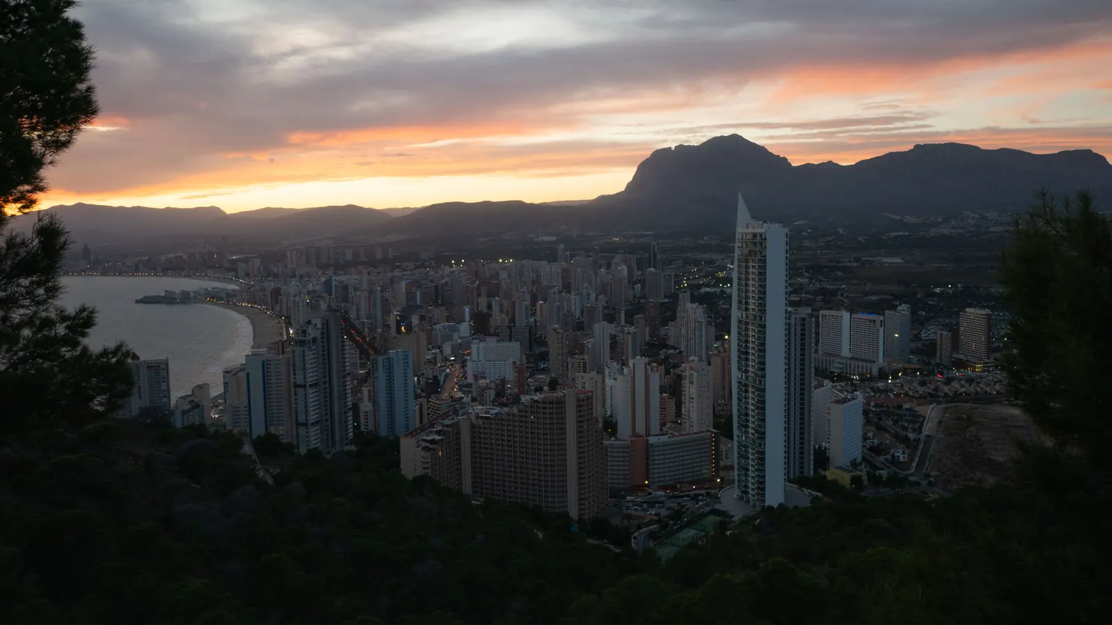Benidorm's tower blocks and the bay under a dusk sky, with a mountain ridge behind the town